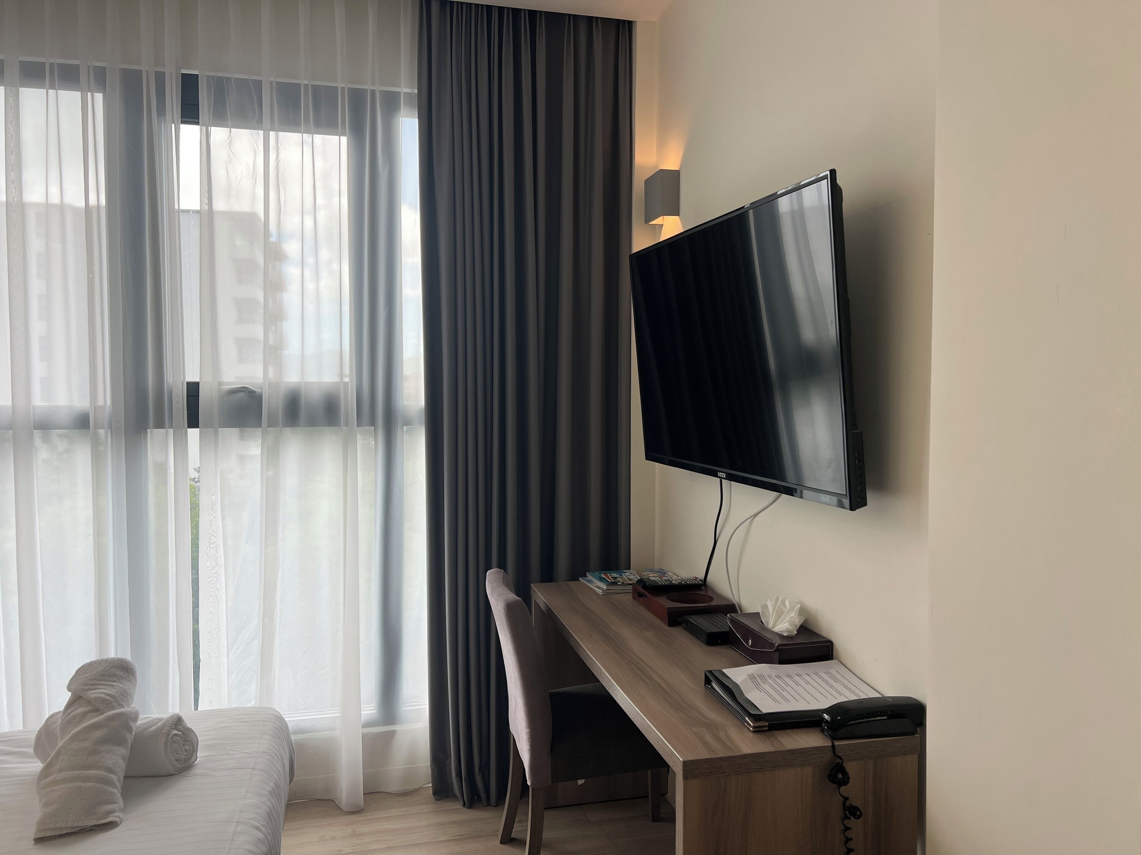 A hotel room with a bed, a desk with a computer, a flat-screen TV, and a window with sheer curtains and a dark drape.