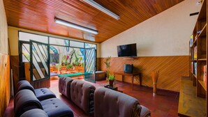 A room with a wooden ceiling, a television on the wall, a sofa, a small table with a plant, and a view through a glass door. - La Quinta - Hostel (Bogotá)