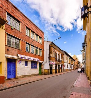 A street with buildings, including a brick building with a blue door and a white building with a closed shutter. - La Quinta - Hostel (Bogotá)