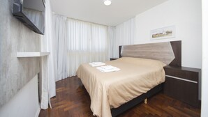 Ein Schlafzimmer mit einem Bett, einem an der Wand montierten Fernseher, einem Holznachttisch und einem Fenster mit Vorhängen.