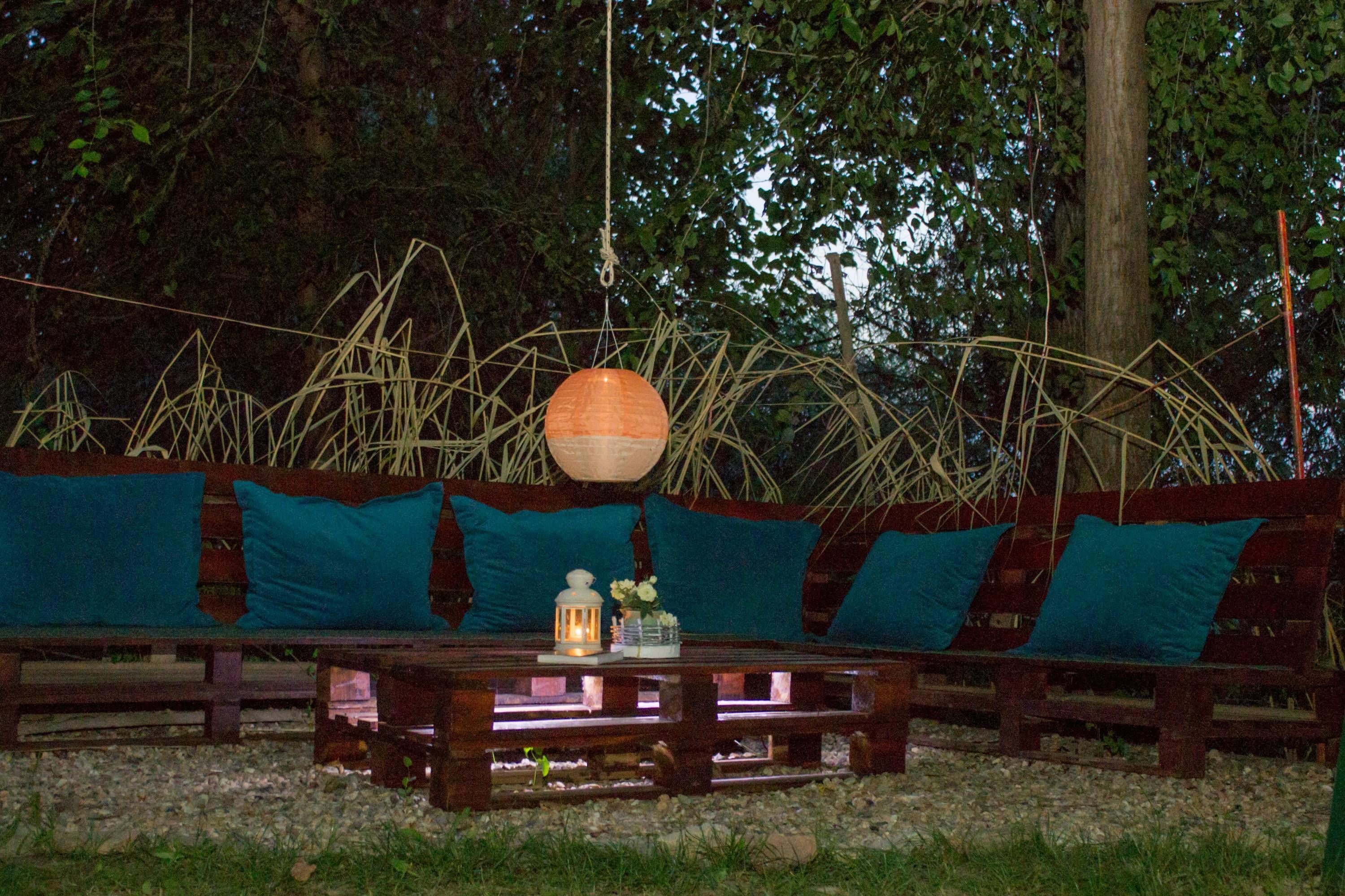Eine hölzerne Bank mit türkisblauen Kissen und einem Laternen auf einem Kiesweg.