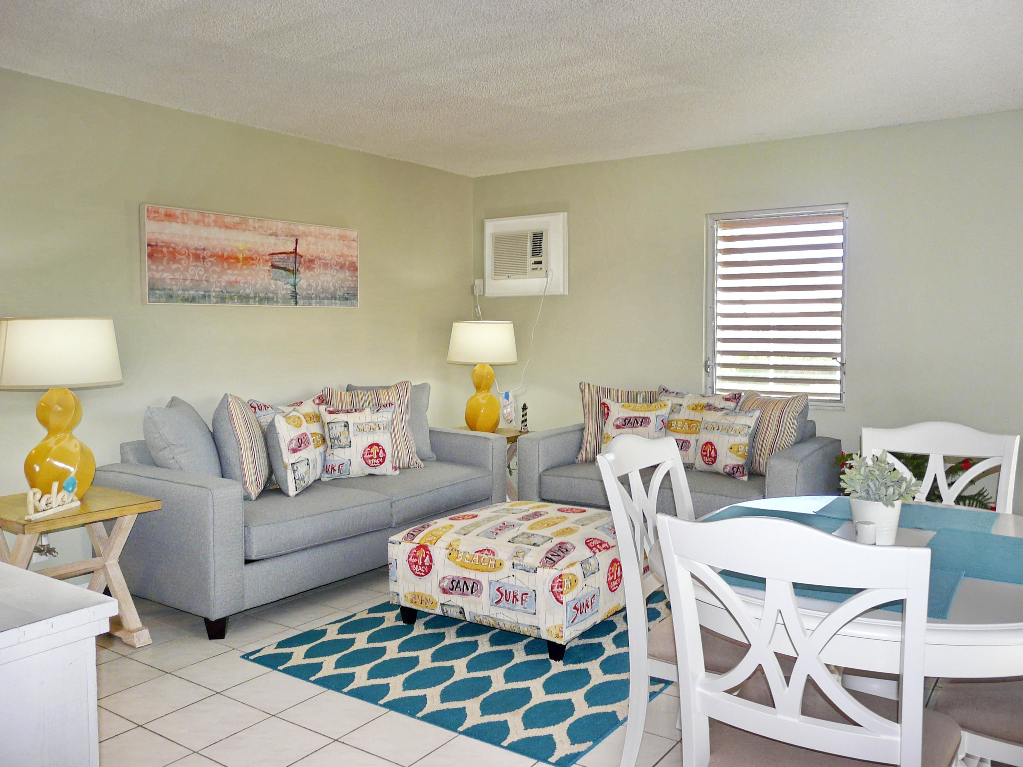 A living room with a grey sofa, a matching ottoman, and a white dining table with chairs. There are two lamps on side tables, a patterned rug, and a beach-themed cushion on the sofa.