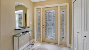 A hallway with a beige wall, a glass console table, a door with decorative metalwork, and a white door to the right. - 601 Lewis Court (Marco Island)