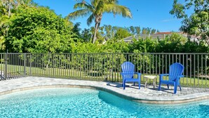 A swimming pool with two blue chairs and a small table, surrounded by a metal fence and lush greenery. - 601 Lewis Court (Marco Island)
