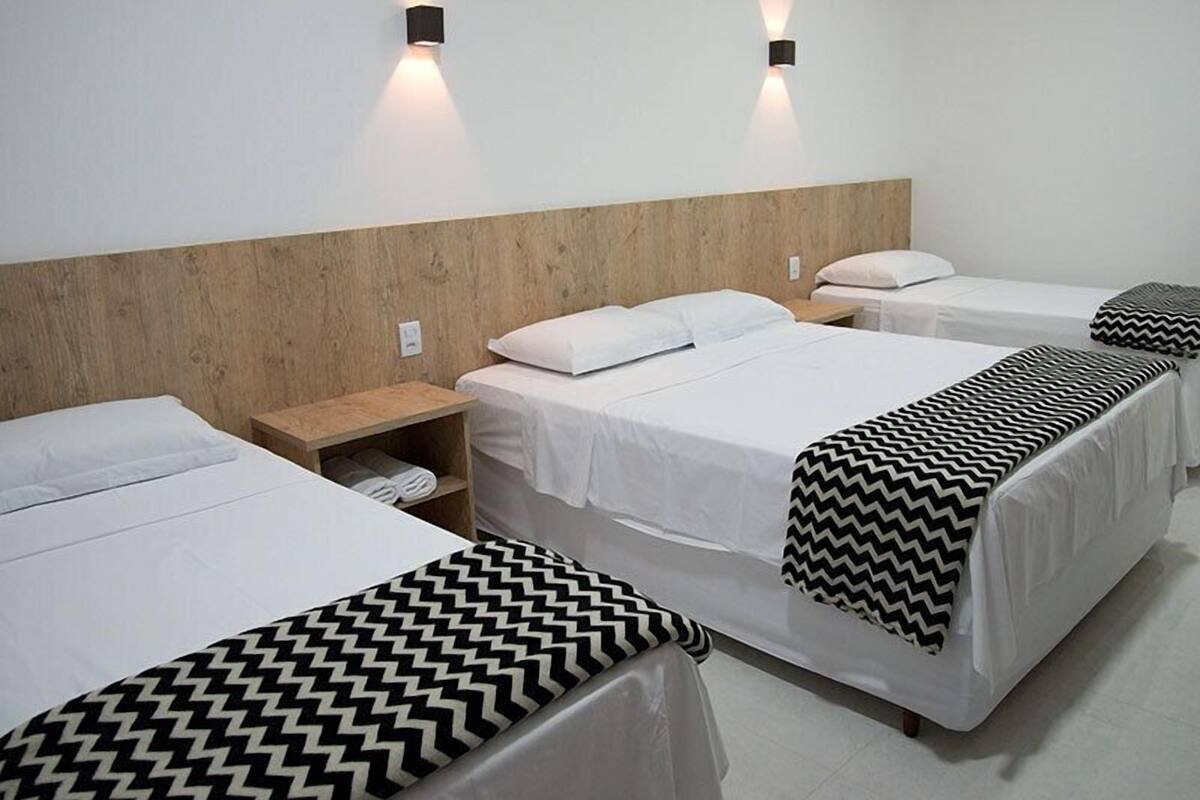 A hotel room with three beds, each with white bedding and black and white patterned throw pillows. There are two wall-mounted light fixtures on either side of the headboard.