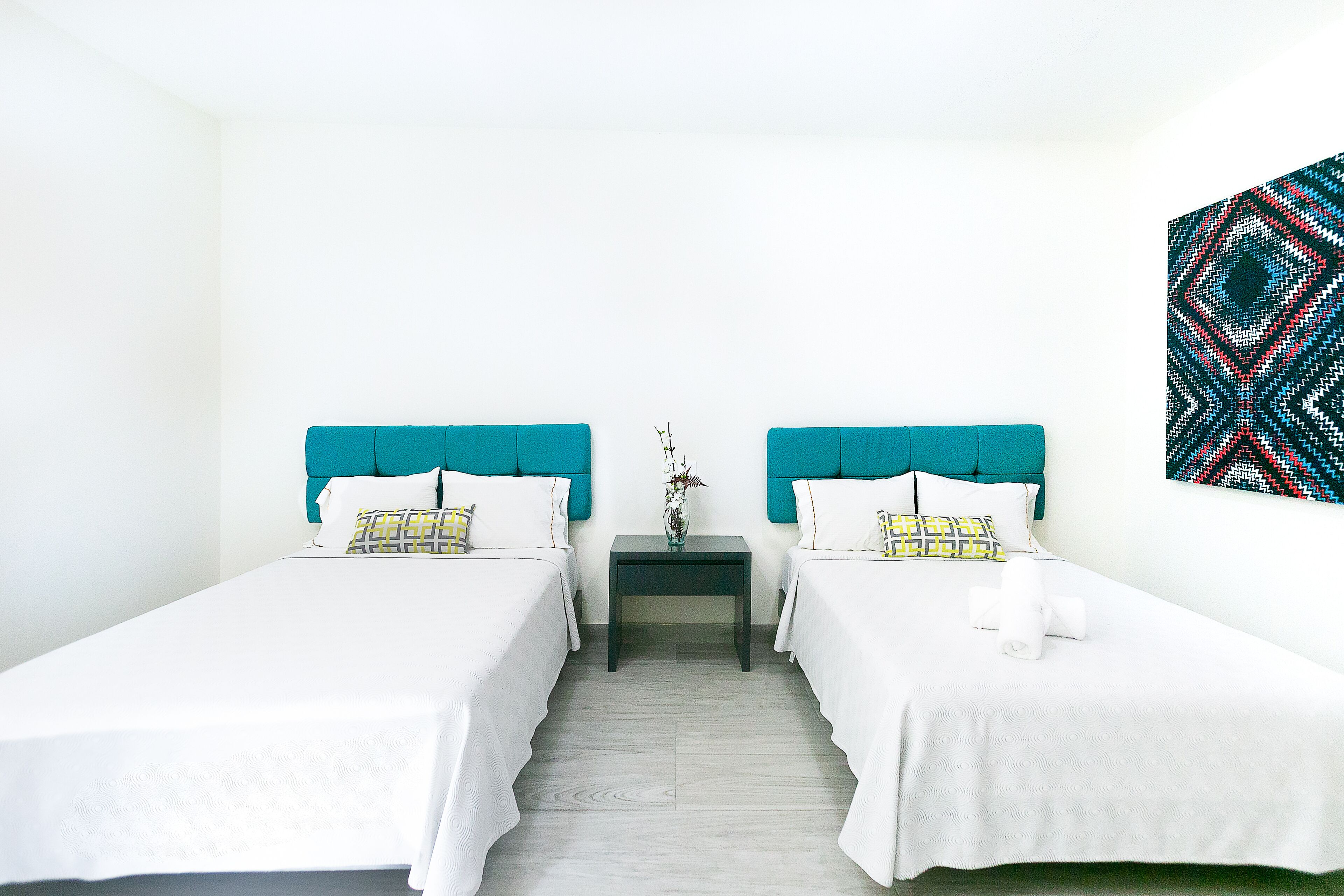 Une chambre d’hôtel avec deux lits simples, chacun doté d’une tête de lit de couleur turquoise, de draps blancs et d’une petite table de chevet sur laquelle est posé un vase.
