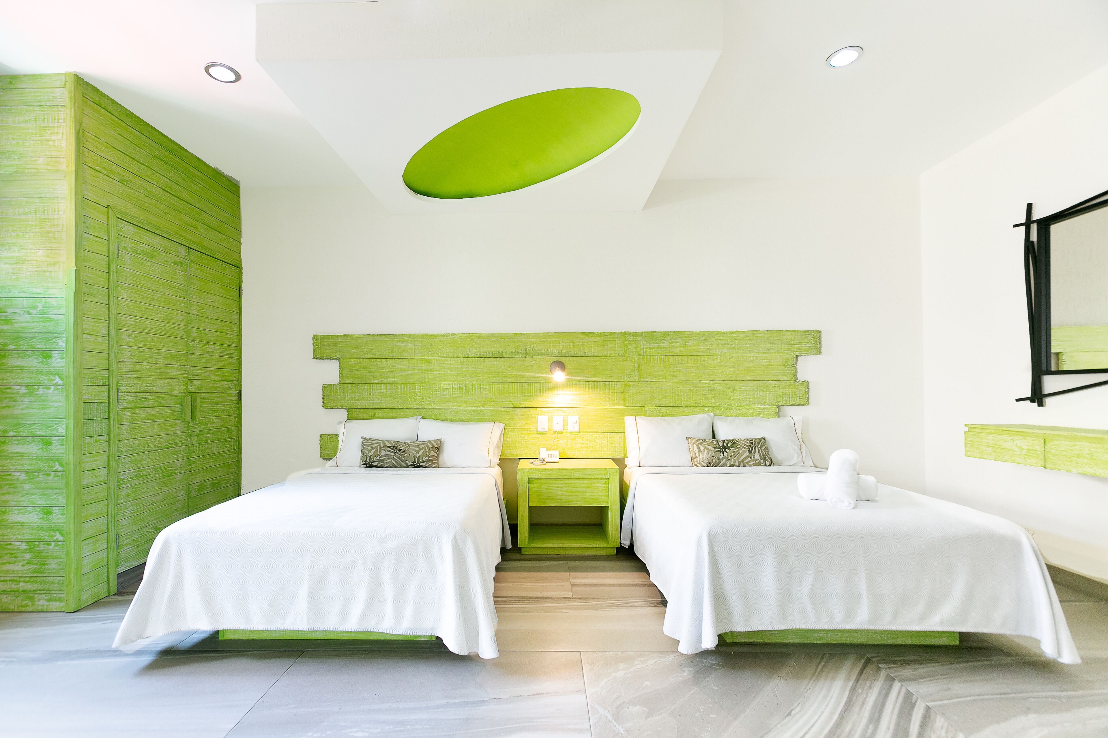 Quarto de hotel com duas camas, parede verde decorativa, luminária no teto e espelho na parede.