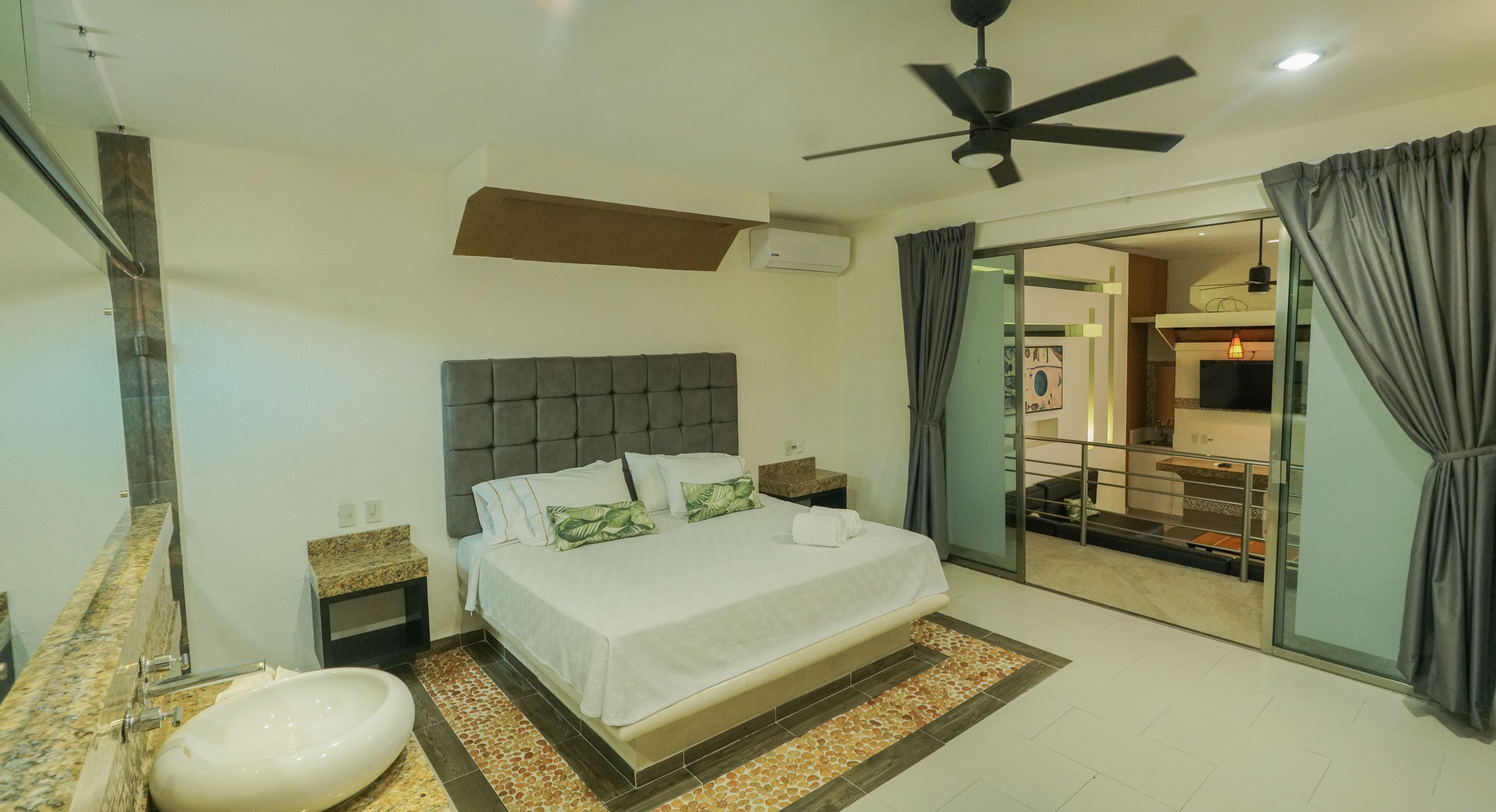 A bedroom with a bed, a ceiling fan, a bathroom with a sink, and a sliding glass door leading to a balcony.