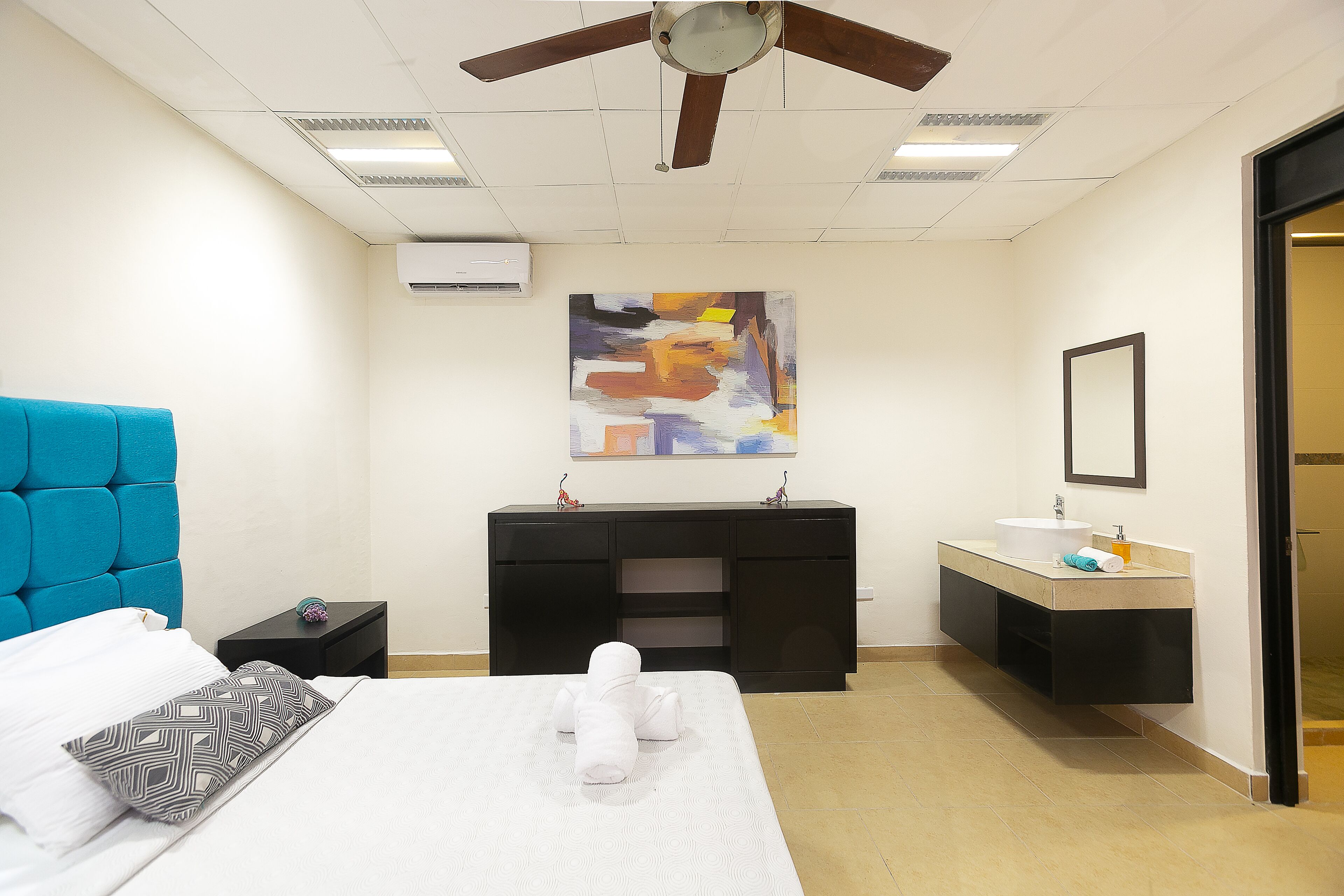 Une chambre d’hôtel comprenant un lit, une table de chevet, une console noire, une salle de bain avec un lavabo et un miroir, et un ventilateur de plafond.