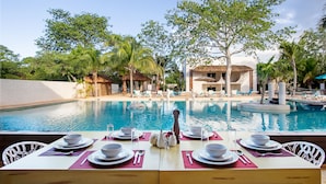 Comedor con mesas dispuestas para una comida, con vista a una piscina con sillas de descanso y palmeras.