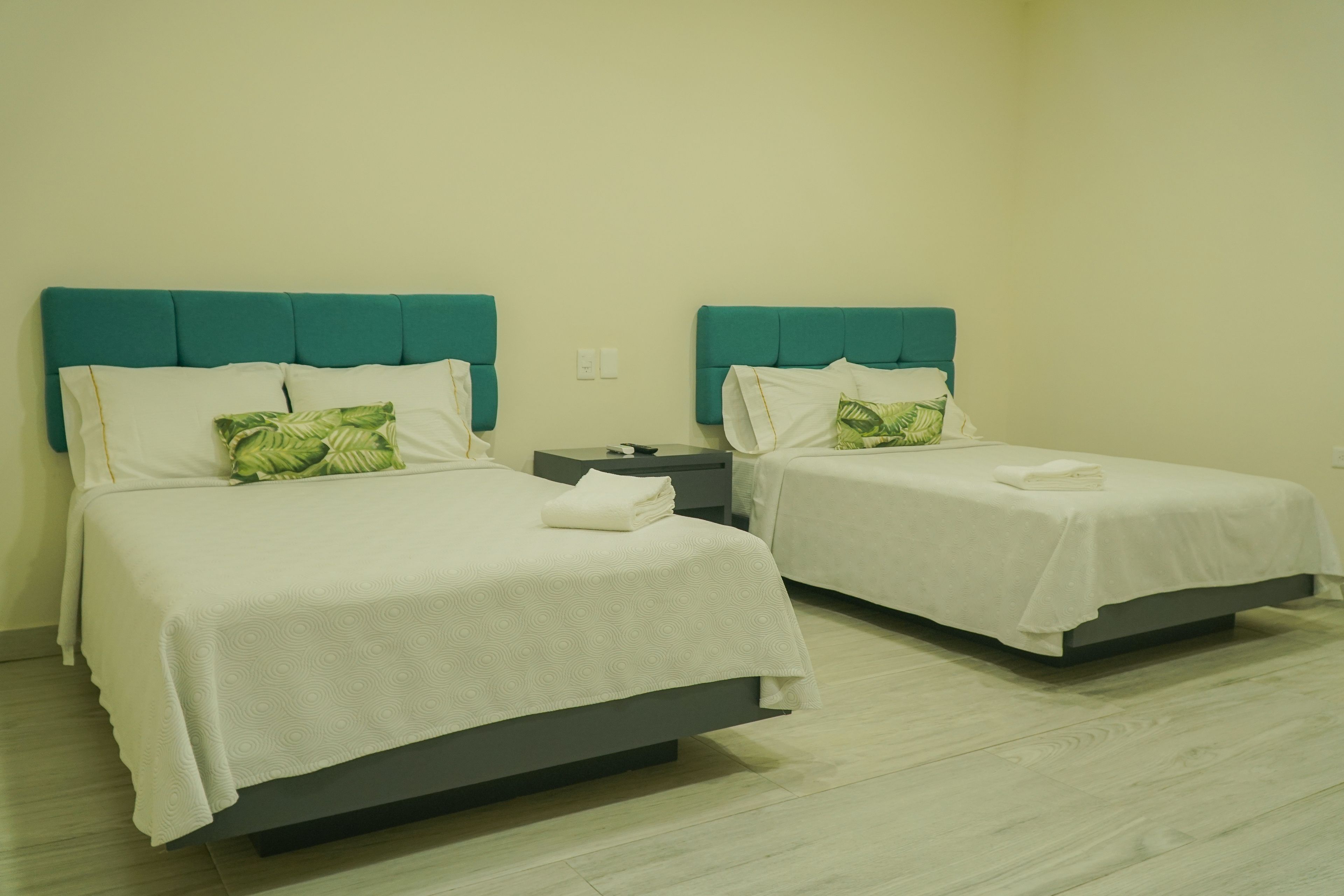 Dos camas individuales con cabeceros color turquesa y ropa de cama blanca, una mesita de noche pequeña y un piso de madera de color claro.