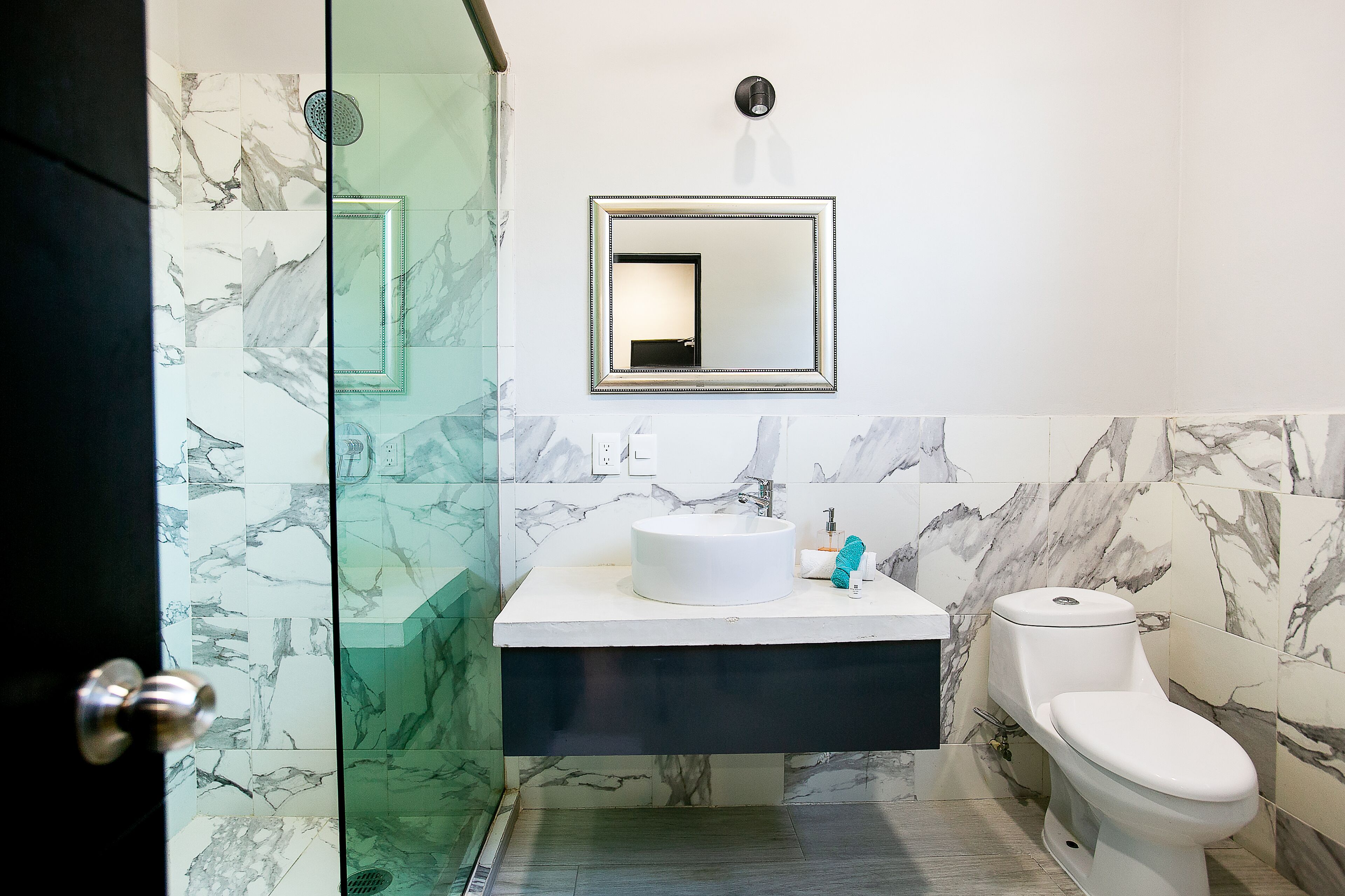 Un baño con pared de azulejos de mármol, un lavamanos blanco, un inodoro y una cabina de ducha de vidrio.
