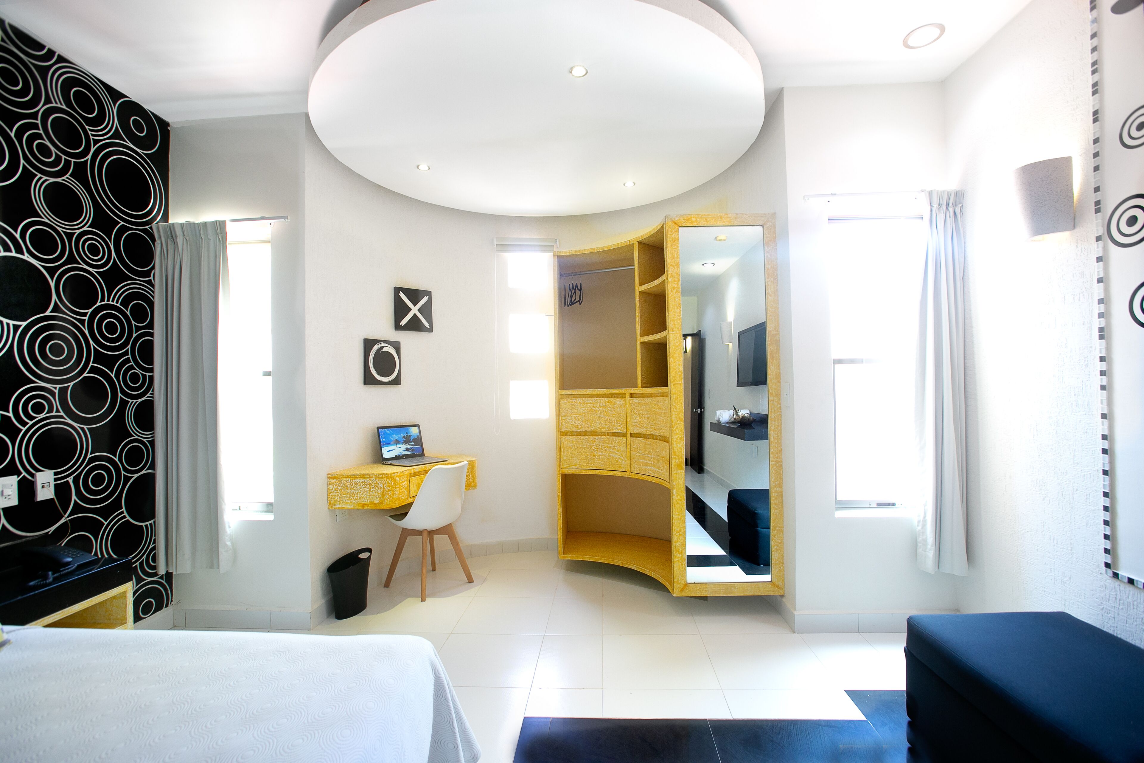 A modern hotel room with a bed, a desk with a computer, a wardrobe, and a bathroom visible through an open door.