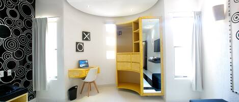 A modern hotel room with a bed, a desk with a computer, a wardrobe, and a bathroom visible through an open door.
