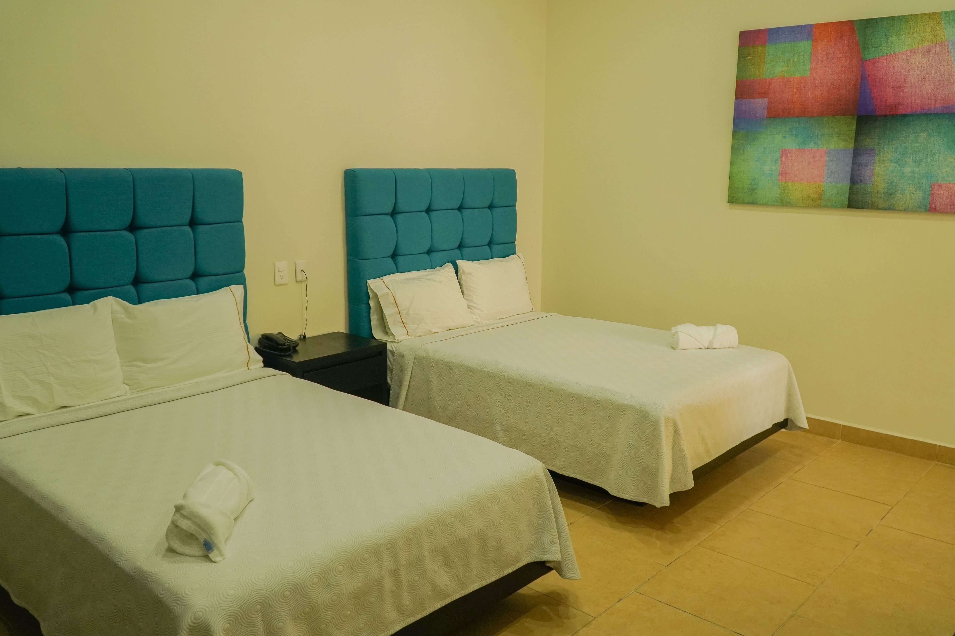 A hotel room with two beds, each with a teal headboard and white bedding. There is a colorful abstract painting on the wall.