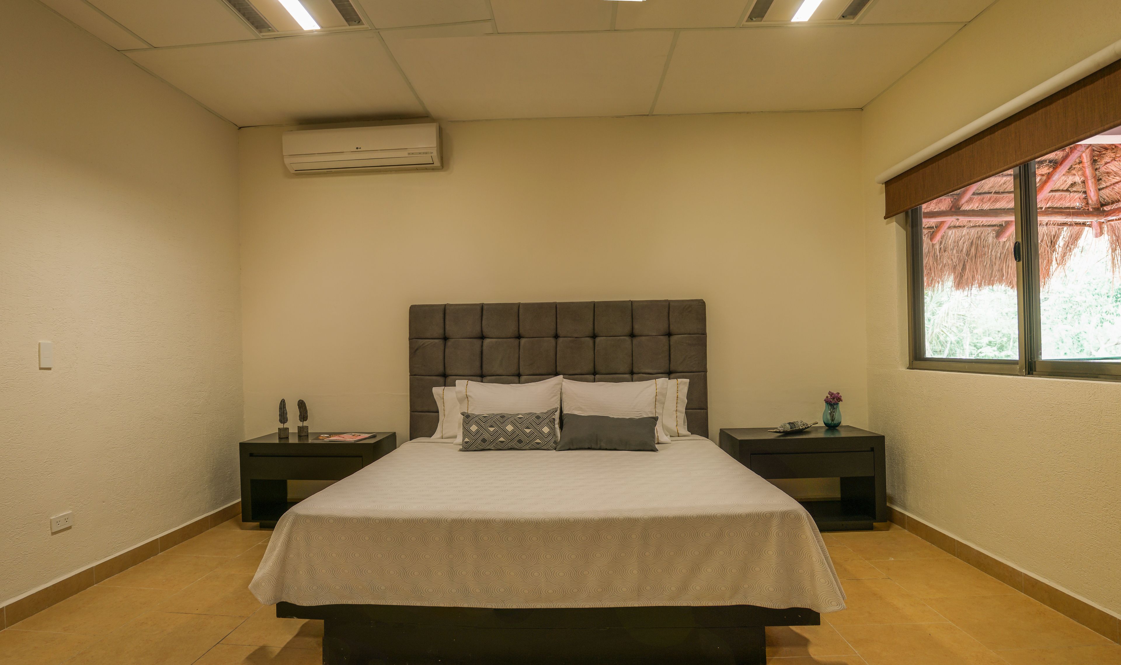 Un dormitorio con una cama grande, dos mesitas de noche, un ventanal con vistas y un aire acondicionado.