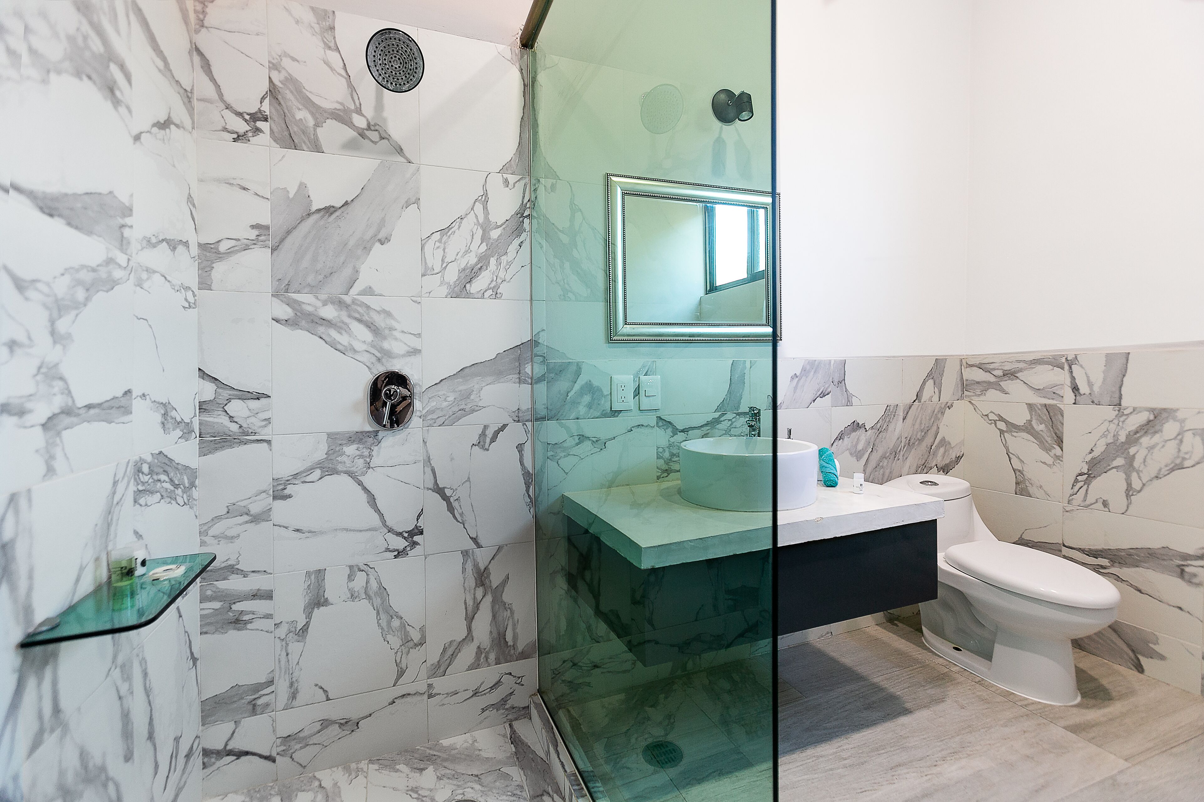 Ein Badezimmer mit Marmorfliesen, einer Glasdusche, einem Waschbecken mit Spiegel und einer Toilette.