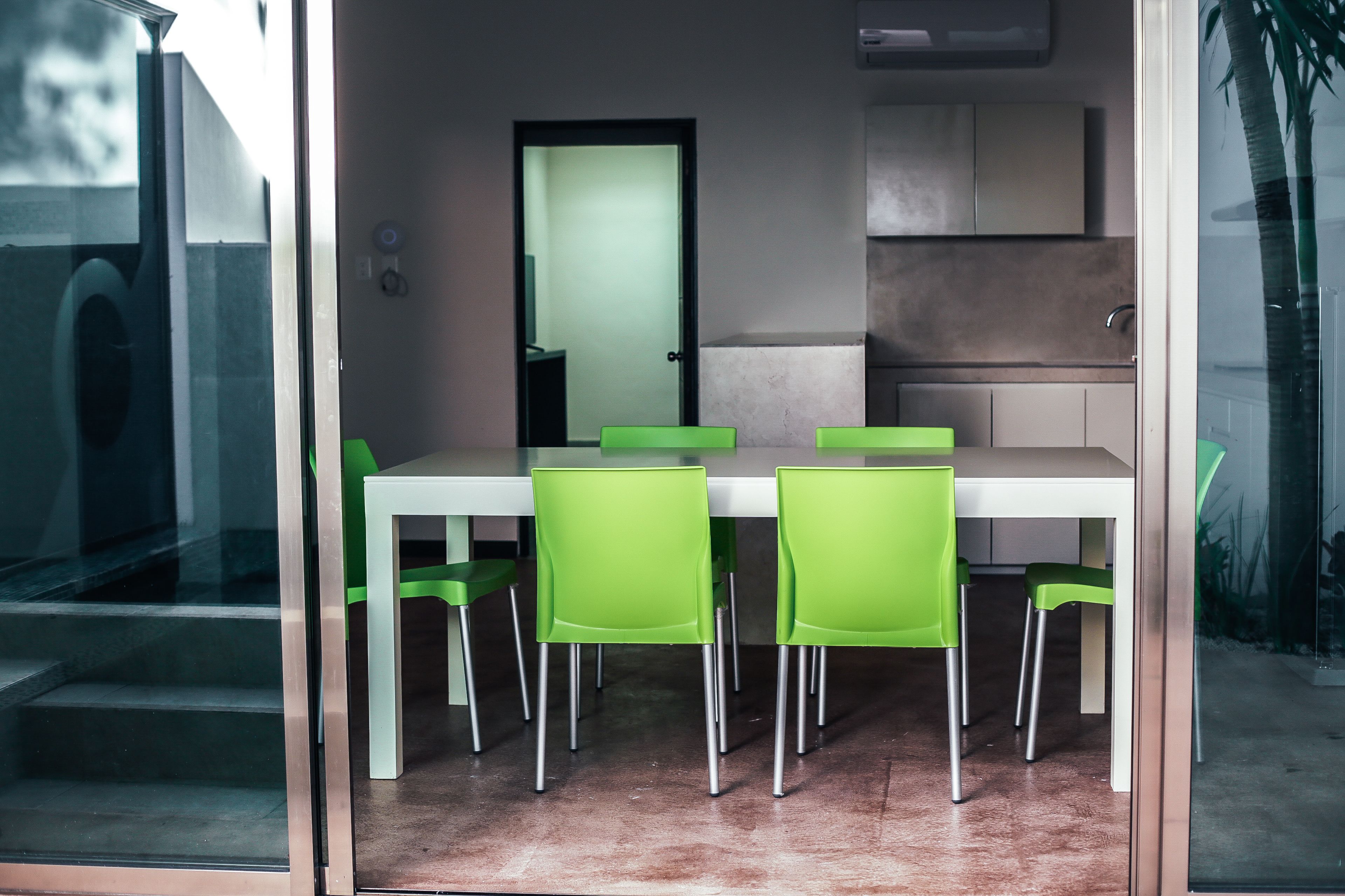 A modern dining area with a white table and green chairs, a glass partition, and a kitchen area in the background.