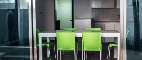 A modern dining area with a white table and green chairs, a glass partition, and a kitchen area in the background.