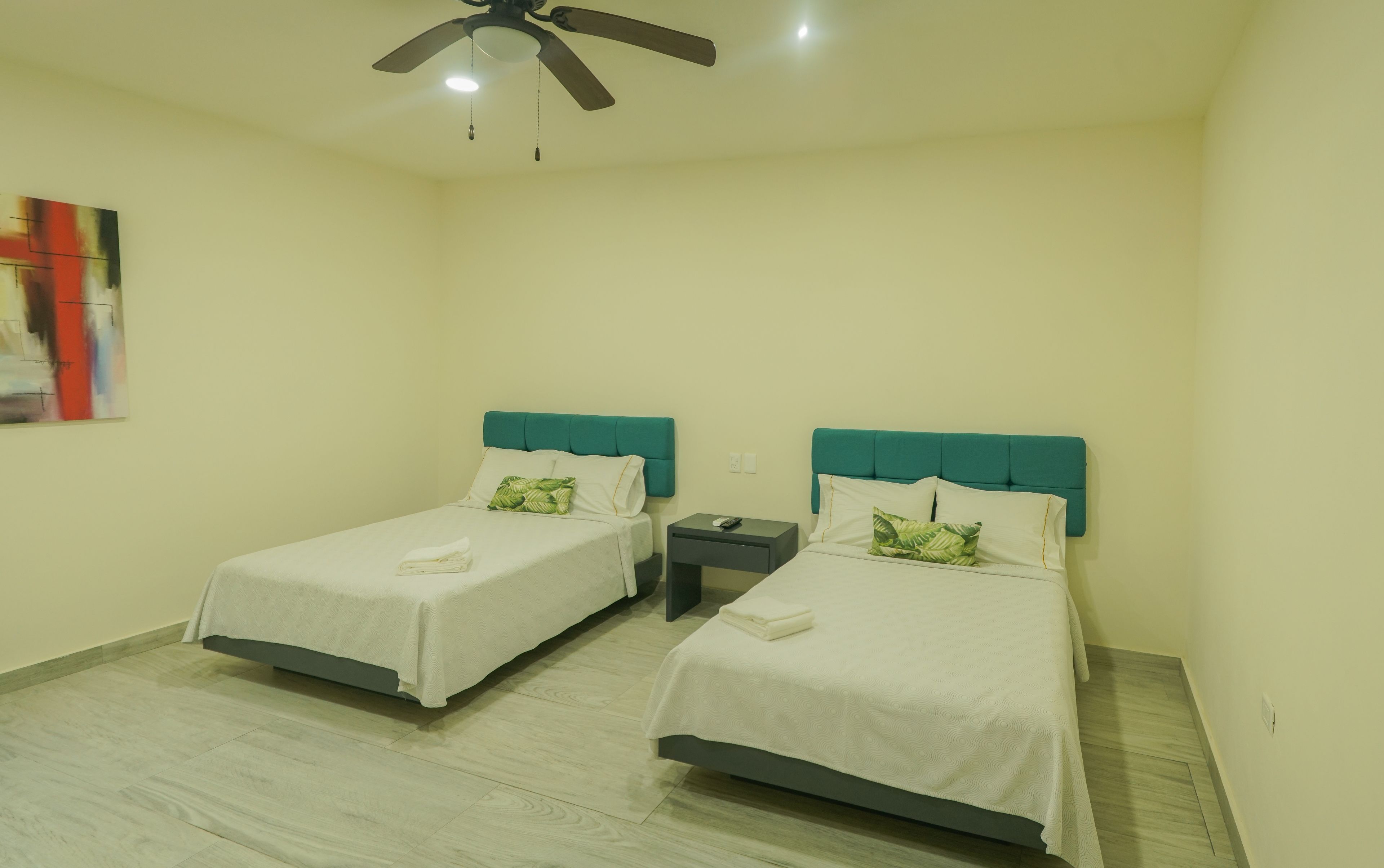 Habitación de hotel con dos camas, cada una con cabecera color aguamarina, ropa de cama blanca y un control remoto en la mesita de noche.