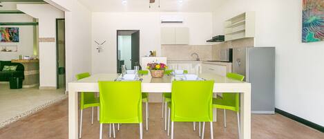 A dining area with a white table and green chairs, a ceiling fan, and a kitchen in the background.