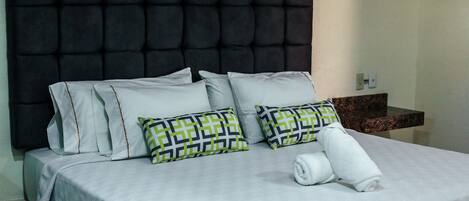 Cama bem arrumada com cabeceira estofada, roupa de cama branca e almofadas estampadas.