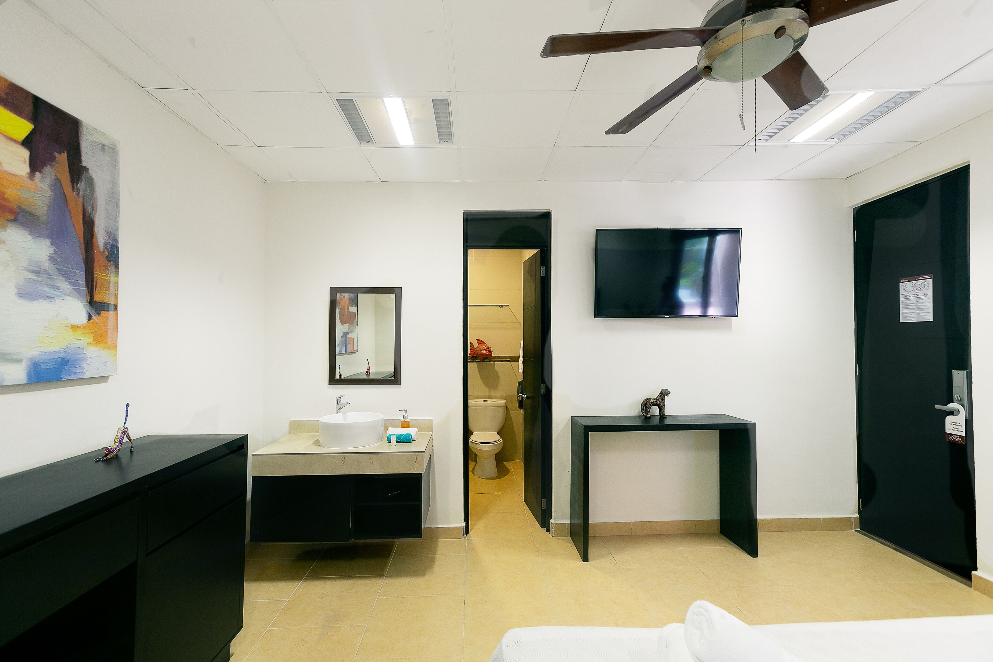 A hotel room with a bed, a desk, a TV, a bathroom with a sink and toilet, and a ceiling fan.