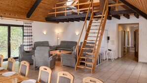 Amplia habitación estilo loft con techo de madera, cama en entrepiso, comedor con sillas y mesa, y sala de estar con sofás y una mesa de centro.