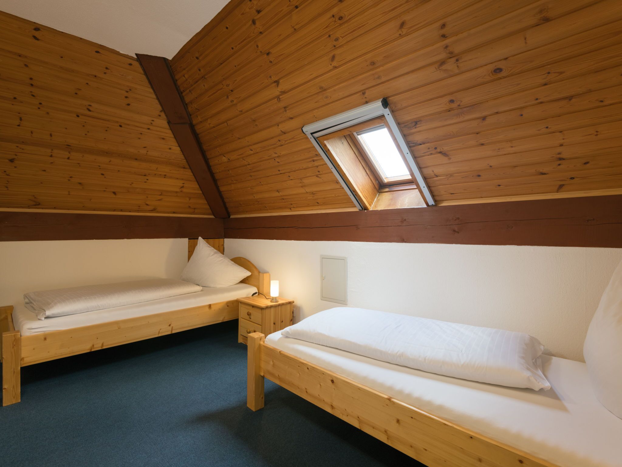 Ein gemütliches Zimmer mit zwei Holzbett, einem kleinen Nachttisch mit Lampe und einem Oberlicht.