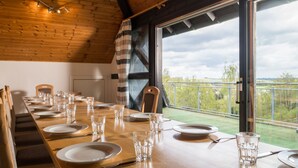 Comedor con una larga mesa de madera dispuesta para una comida, ventanas amplias con vistas al exterior y techo de madera.