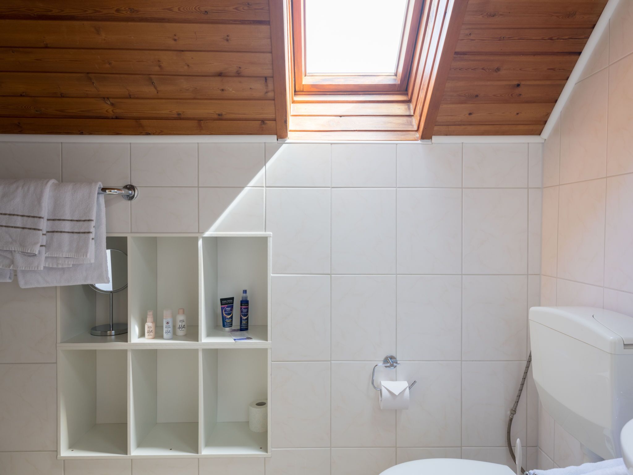 Un baño con tragaluz, azulejos blancos, un inodoro y un estante con artículos de aseo.