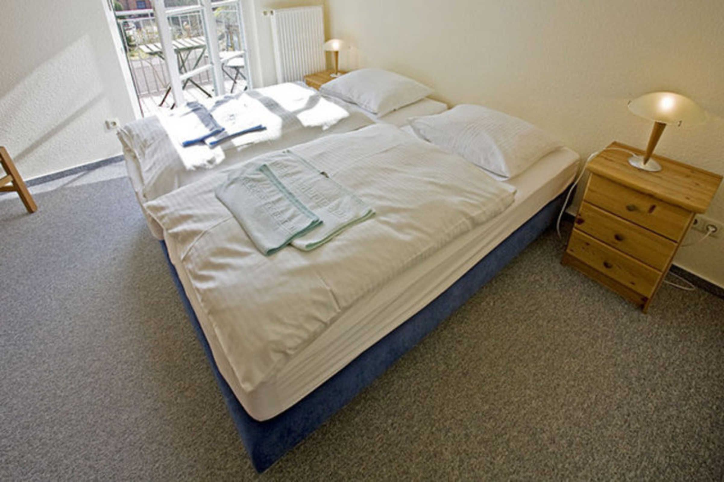 A double bed with a blue mattress, two bedside tables with lamps, a wooden nightstand, and a view of the outside balcony.
