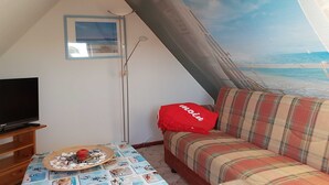 Ein gemütliches Zimmer mit Fernseher, einem Tisch mit dekorativer Platte, einer Couch mit kariertem Überwurf und einem großen Fenster mit Blick auf den Strand.