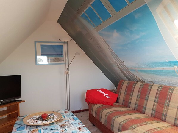 A cozy room with a television, a table with a decorative plate, a sofa with a plaid blanket, and a large window with a beach view.