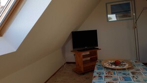 Ein Zimmer mit schrägem Dach, einem Fernseher auf einem Holzständer, einem Tisch mit gemustertem Tischläufer und einem kleinen gerahmten Bild an der Wand.
