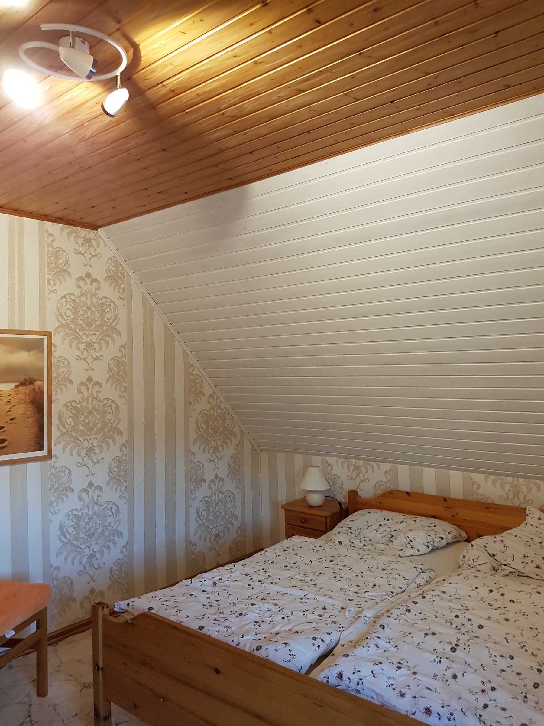 Quarto com teto inclinado, cama com roupa de cama floral, mesa de cabeceira com abajur e um quadro na parede.