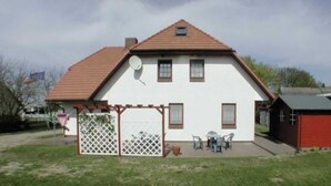 Ein zweistöckiges Haus mit geneigtem Dach, einem eingezäunten Patio mit Gartenmöbeln und einem kleinen Schuppen zur Rechten.