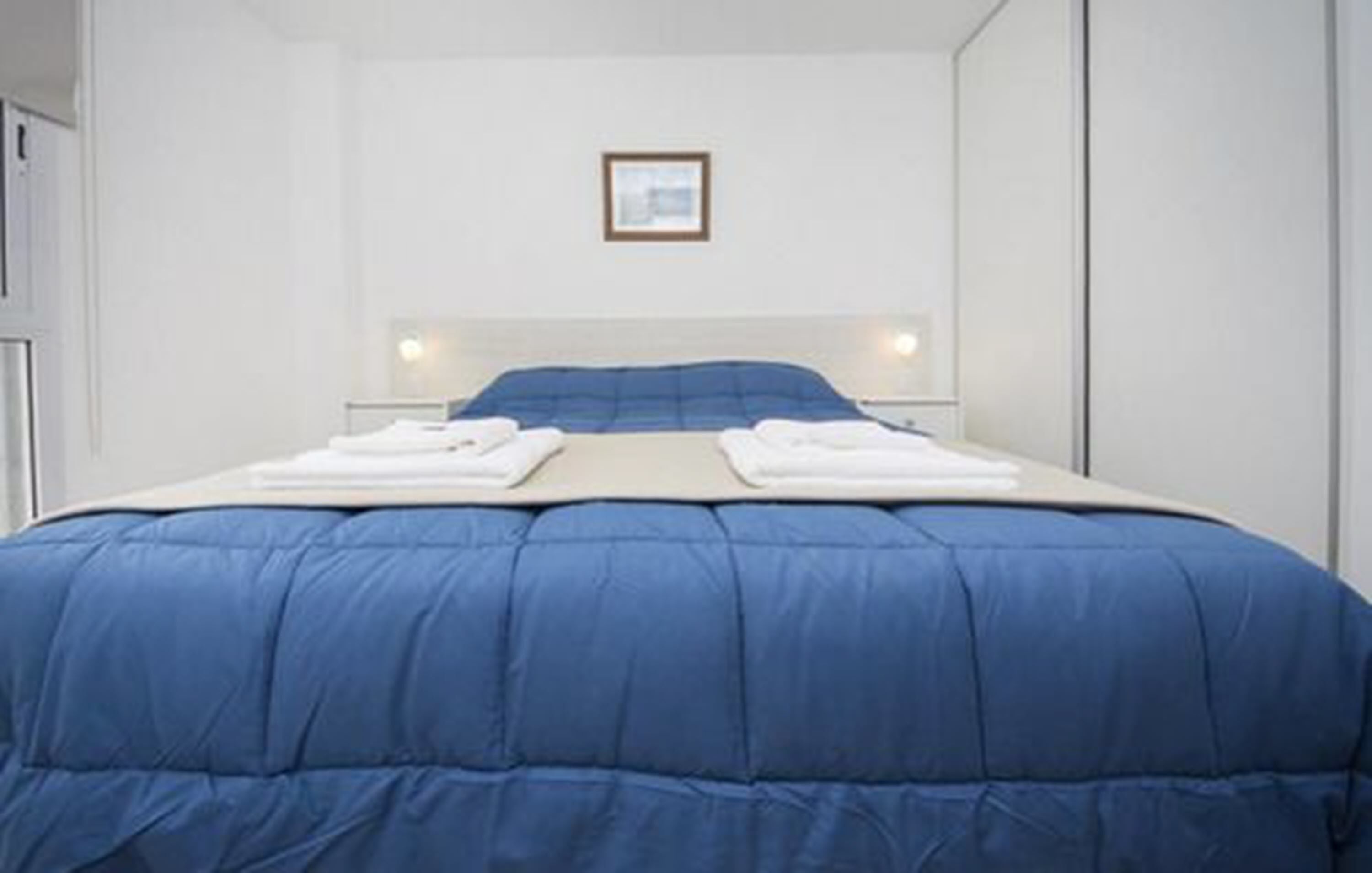 A neatly made bed with a blue comforter and white linens, a framed picture on the wall, and a simple, clean room design.