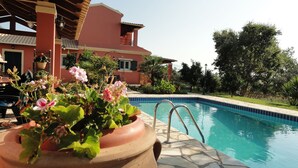 A swimming pool with a tiled edge, surrounded by a garden with potted plants and a building with a red facade. - Villa Thea (Corfu)