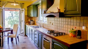 A kitchen with green cabinets, a tiled backsplash, a gas stove, and a dining area with a chair and table. - Villa Thea (Corfu)