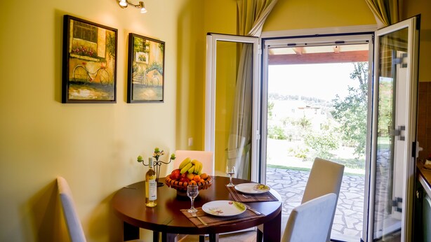 A dining table with a fruit bowl, wine glasses, and a bottle of wine. There are two framed paintings on the wall and a sliding glass door leading to an outdoor area. - Villa Thea (Corfu)