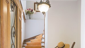 Un escalier en bois, avec un luminaire au-dessus, et un porte-chapeaux sur lequel sont posés un chapeau et un tissu rose.
