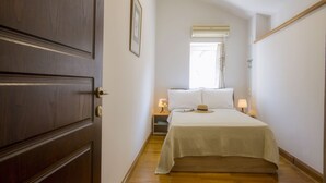 Une chambre d’hôtel étroite avec une porte en bois, un lit recouvert d’une couette blanche, une table de chevet avec une lampe et une fenêtre avec un rideau.