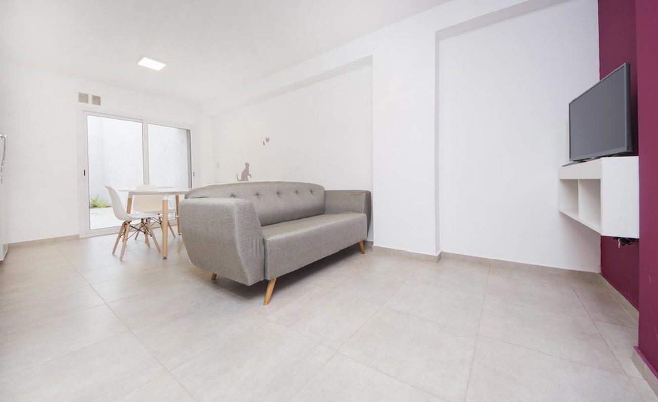 A modern living room with a grey sofa, a dining table with chairs, and a flat-screen TV mounted on a white wall.