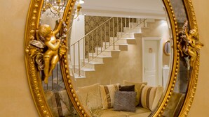 An ornate gold-framed mirror reflecting a staircase and a living room with a sofa and cushions. - Suite Maison (Corfu)