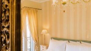 A bedroom with a large bed, a chandelier, a nightstand, and a window with curtains. - Suite Maison (Corfu)