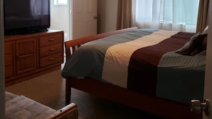 A bedroom with a bed, a ceiling fan, a TV on a wooden cabinet, and a window with curtains.