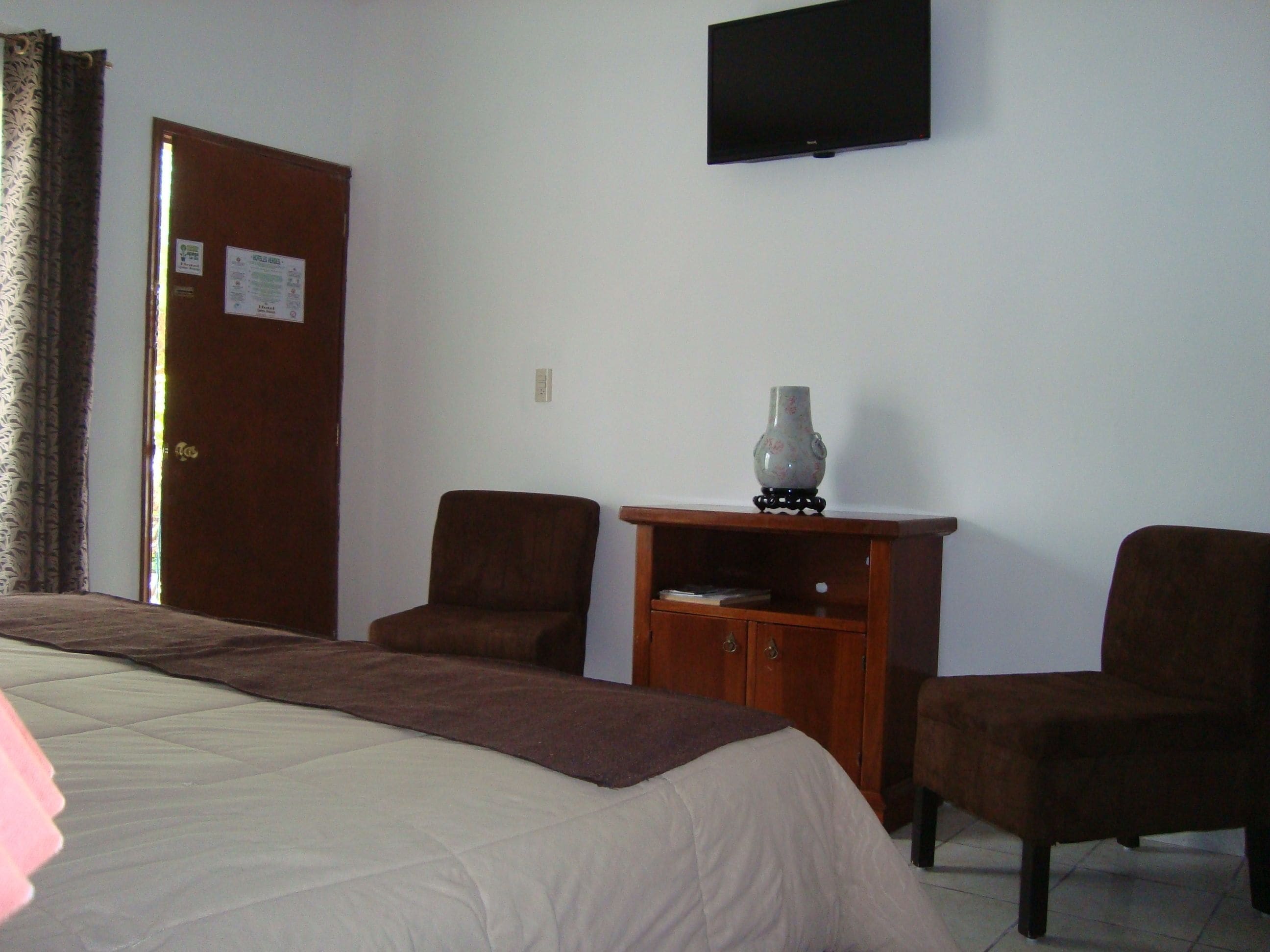 Une chambre d’hôtel comprenant un lit, deux fauteuils, une armoire en bois, un vase, un téléviseur à écran plat et une porte sur laquelle est affichée une note.