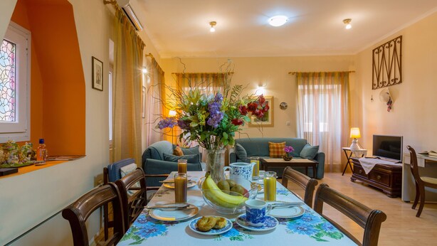 A dining area with a table set for breakfast, a vase with flowers, and a living room in the background. - Heart of the old town (Corfu)