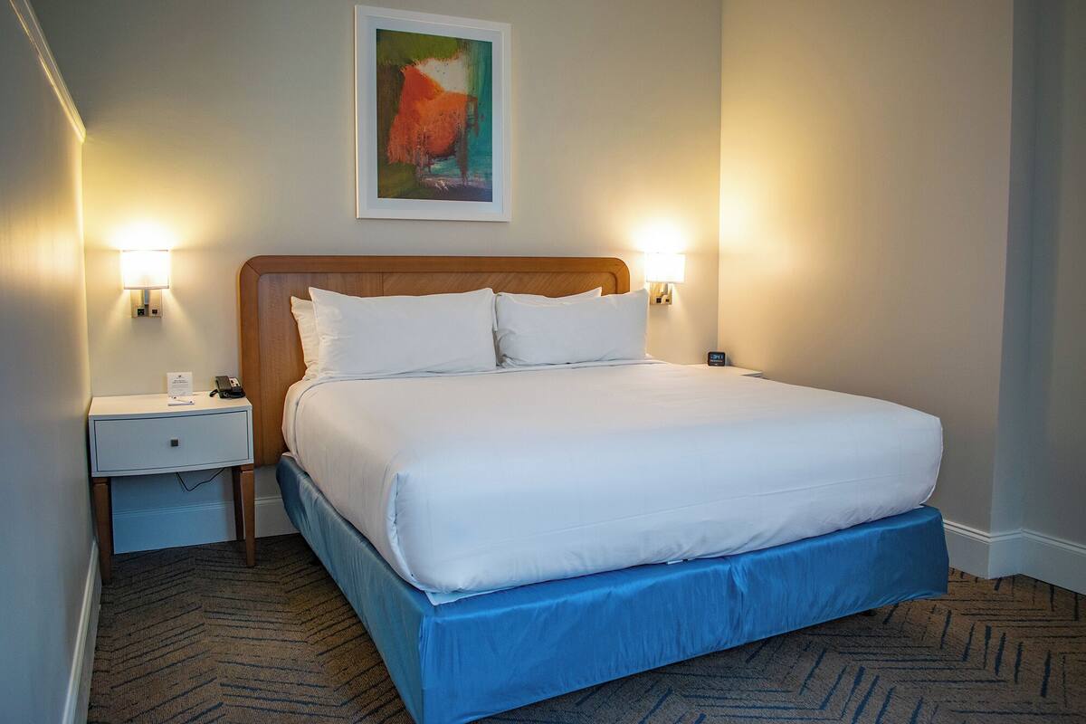 A hotel room with a large bed, two bedside tables, a nightstand, a painting on the wall, and two wall-mounted lamps.